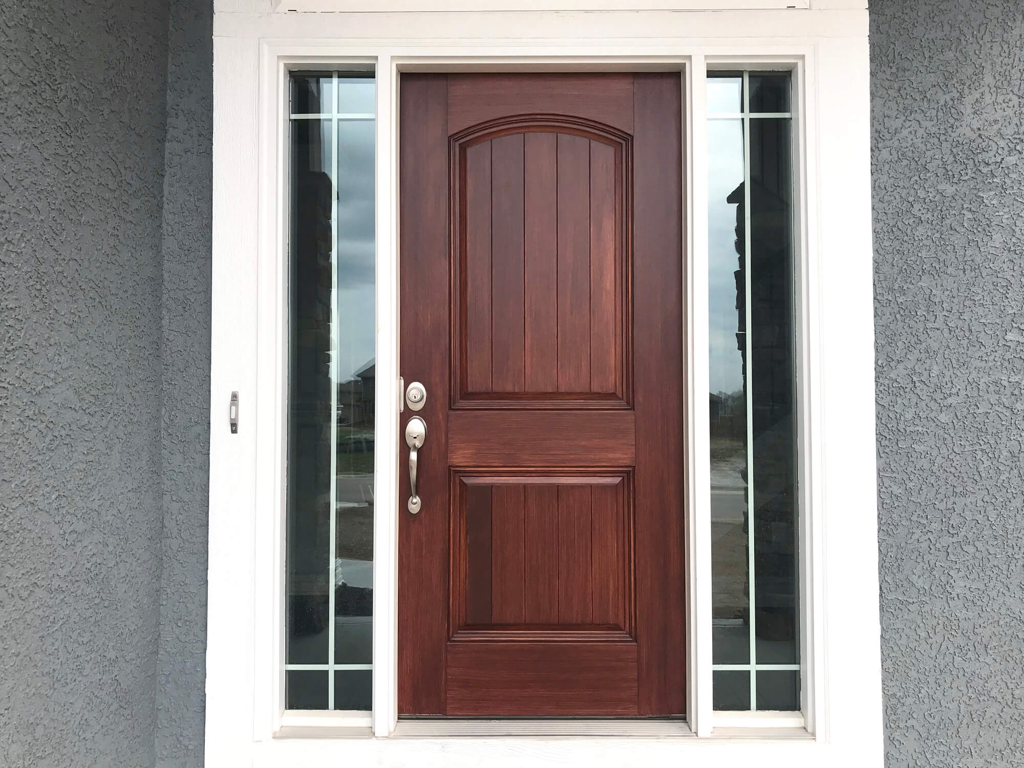 Rich mahogany two-panel arched front door with dark glass sidelights on grey stucco house