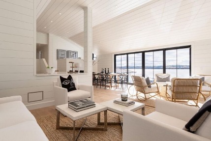 A photo of the inside of an open-concept living area in a home that has white shiplap walls, and black-framed lift and slide doors that show a view of a lake.